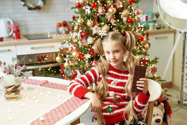 Happy little girl eating chocolate at table in domestic kitchen with Christmas tree