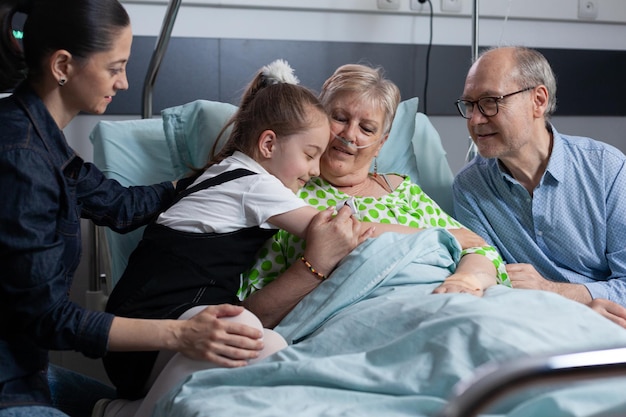 Happy little girl greeting sick grandmother with hug on hospital room visit. Old lady lying on medical bed happily giving hugs to granddaughter at geriatric patients medical clinic.