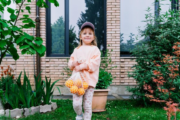 Happy little girl holding oranges in mesh bag in front of house in green garden