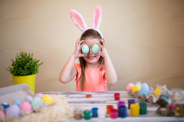 Счастливая маленькая девочка держит пасхальные яйца возле глаз, готовясь к пасхе в кроличьих ушах