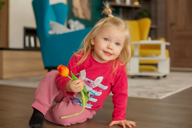 a happy little girl in home clothes is sitting on the floor with a tulip in her hands