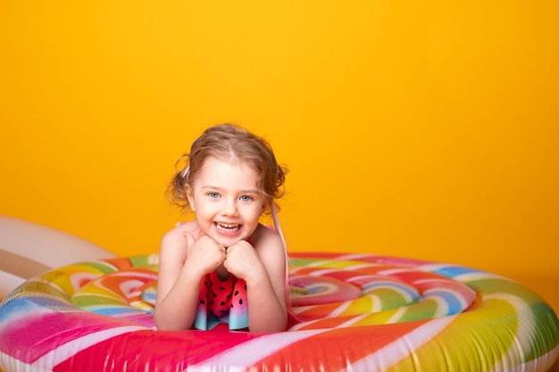 Фото Счастливая маленькая девочка в купальном костюме, лежащем на красочном надувном леденце матраса на желтой поверхности.