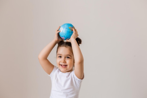 A happy little girl is holding a small globe planet in her hands in a white tshirt on a white