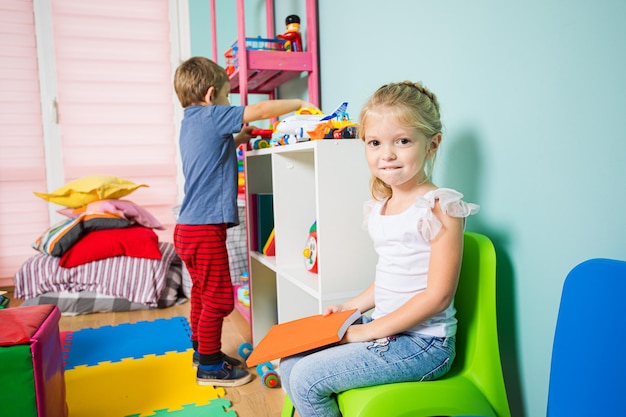 Счастливая маленькая девочка сидит на цветном стуле в детском саду. Она держит в руках книгу, а на заднем плане играет мальчик.