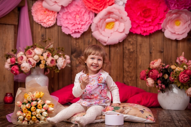 Happy little girl is sitting on pillows on wooden floor, surrounded by flowers .