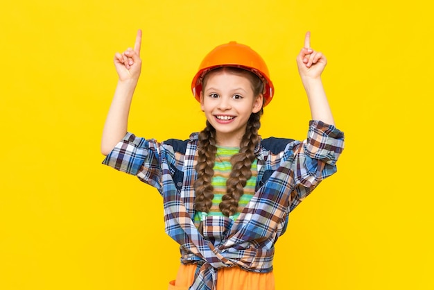 The happy little girl is wearing a construction helmet and a shirt smiling broadly and pointing up The child chooses repairs in the nursery Yellow isolated background