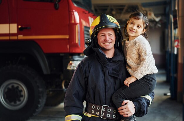Happy little girl is with male firefighter in protective\
uniform