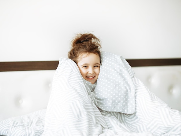 Happy little girl kid covered with blanket sitting on bed looking at camera and laughs