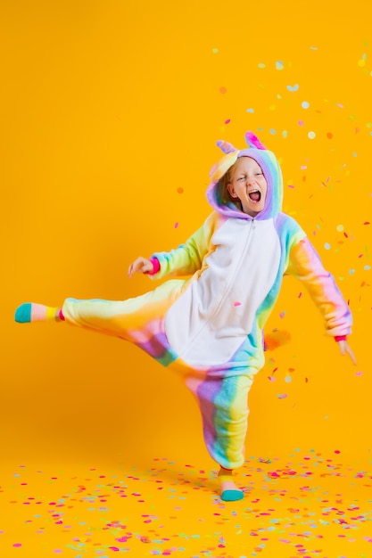 Happy little girl in kigurumi unicorn dancing on a yellow wall among multicolored confetti