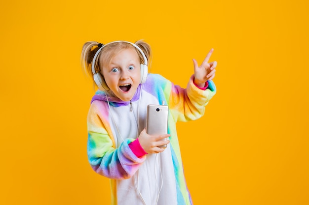 A happy little girl in a kigurumi unicorn listens to music in headphones holding a smartphone