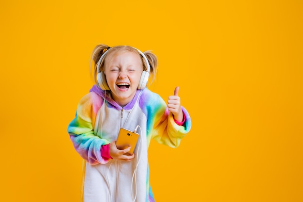 A happy little girl in a kigurumi unicorn listens to music in headphones holding a smartphone