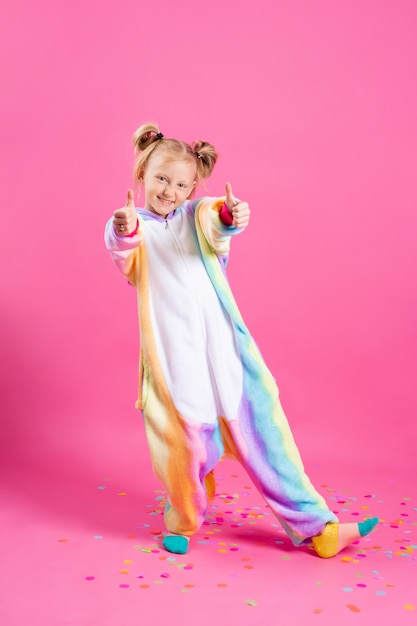 Happy little girl in kigurumi unicorn on a pink wall rejoices in multi-colored confetti