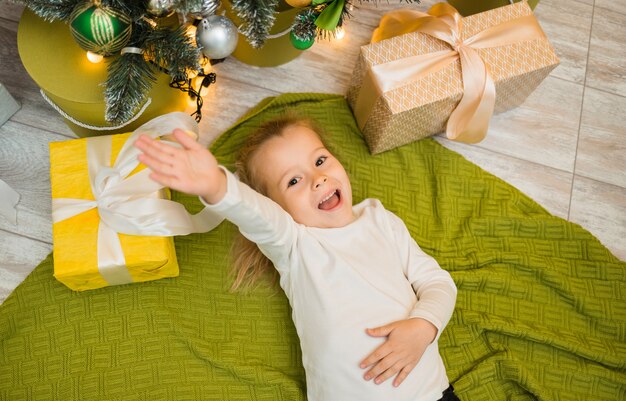 A happy little girl lies on a green knitted blanket under a Christmas tree with gifts and stretches her hand