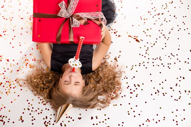 A happy little girl lies on a white background with a gift box and a cap for her birthday.