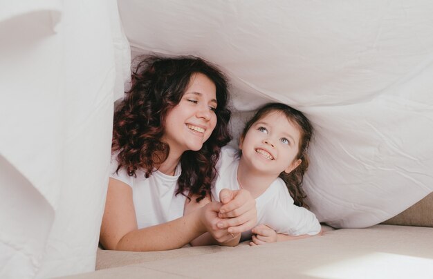 A happy little girl in a light turtleneck hugs her mother under a white sheet. a loving and caring mother