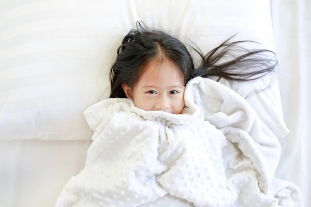 Happy little girl lying on bed with blanket and looking at camera. Top view.