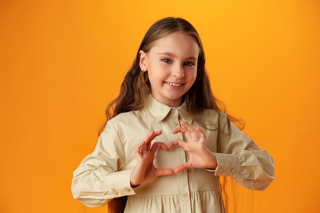 Happy little girl making heart shape by hands on yellow background