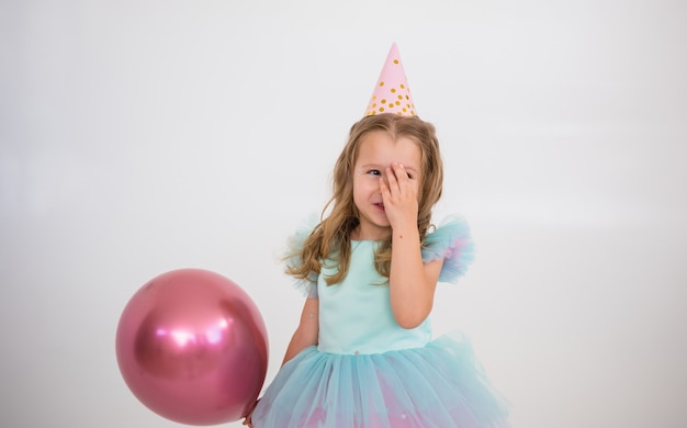 A happy little girl in a paper cap and a smart dress stands with a pink balloon on a white background with a place for text