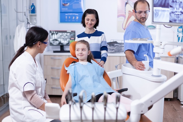 Happy little girl at pediatric dentist wearing dental bib during stomatology examination. Child with her mother during teeth check up with stomatolog sitting on chair.