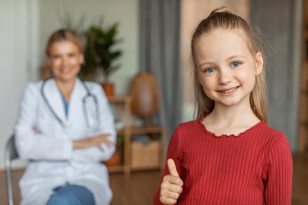 Happy little girl at pediatrician clinic with female doctor on background smiling at camera and showing thumb up