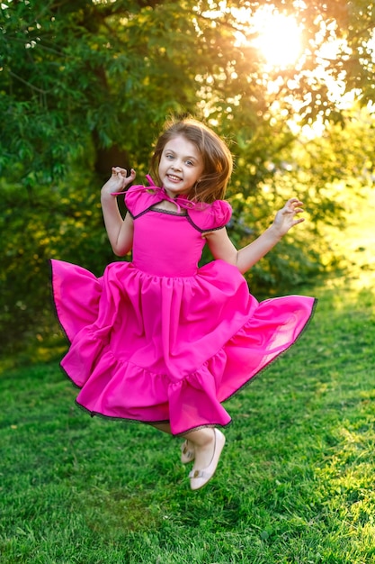 Счастливая маленькая девочка в розовом платье улыбается и прыгает по траве летом на закате