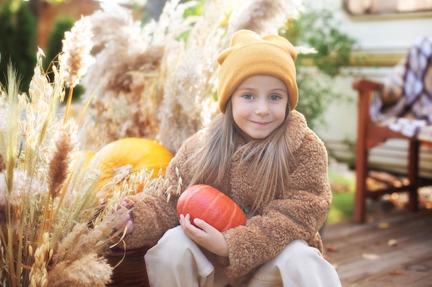 Happy little girl playing with a pumpkin near home in fall garden in cozy fall backyard