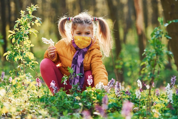 Happy little girl in protective mask sitting in spring forest at daytime