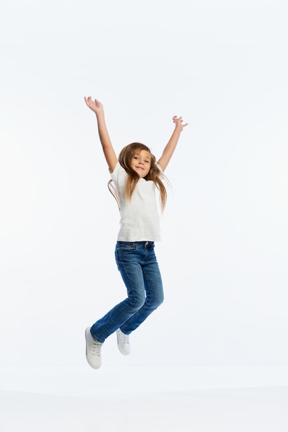 Happy little girl pupil in casual style clothes jumping with rejoice and raising hands up isolated