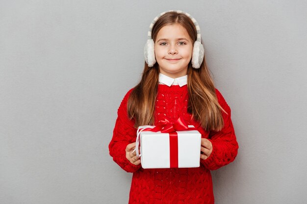 Happy little girl in red sweater and earmuffs with present box
