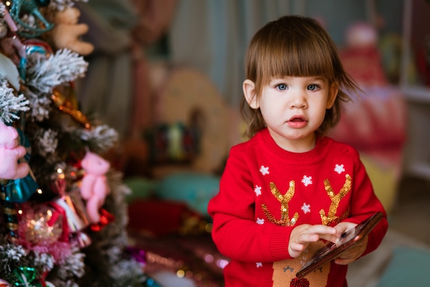 Happy little girl in red sweater joyfully holding phone next to decorated christmas tree on christma...