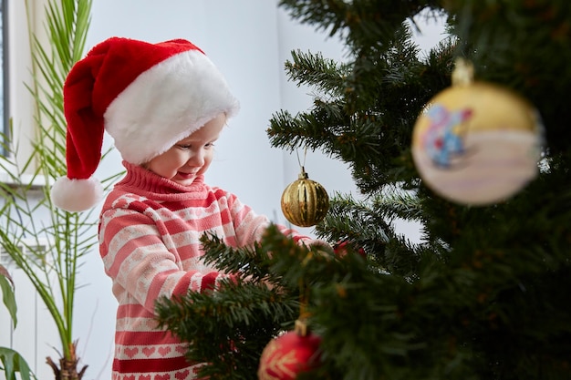 Happy little girl in a Santa Claus hat is decorating the Christmas tree with toys and trinkets. Cute baby is decorating home to celebrate Christmas.
