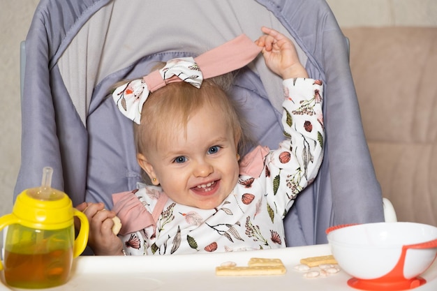 A happy little girl sits in a high chair with a bottle and a plate and looks into the camera smiling