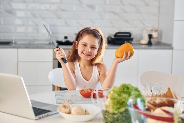 Happy little girl sits on the kitchen with food and laptop on the table