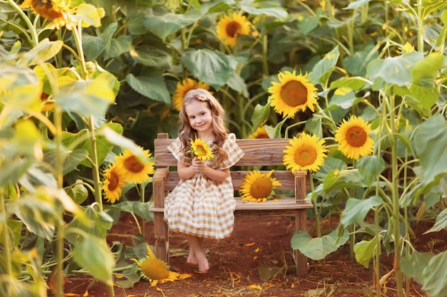 Happy little girl sitting on a bench among blooming sunflowers under the rays of the sunset