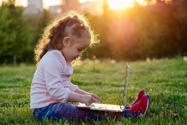 Happy little girl sitting on grass with laptop in park