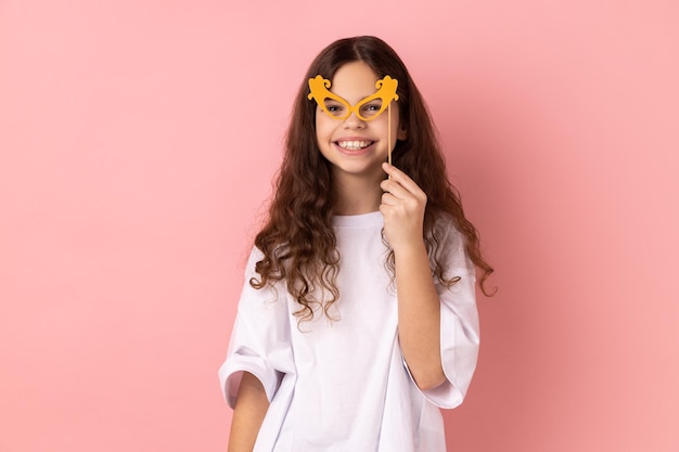 Bambina felice che sorride e tiene gli occhiali di carta sul bastone usando gli oggetti di scena in vacanza divertendosi