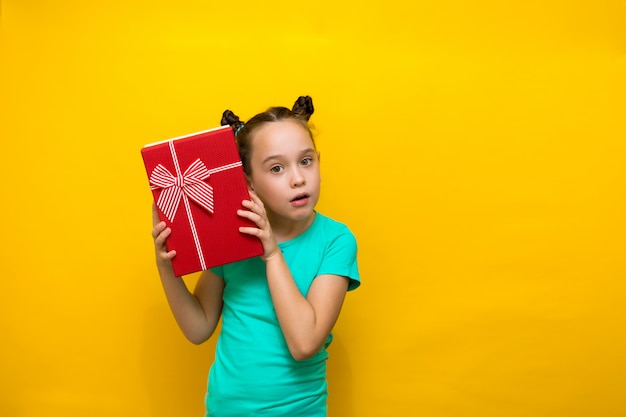 Happy little girl standing isolated over yellow wall will listen in surprise to the box, holding it to her ear