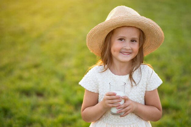 麦わら帽子をかぶった幸せな女の子が芝生の上でグラス一杯の牛乳を飲む