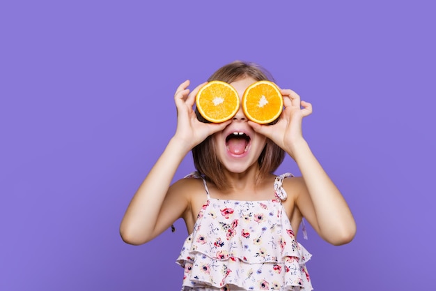 Happy little girl in summer dress and straw hat holding an orange on purple background