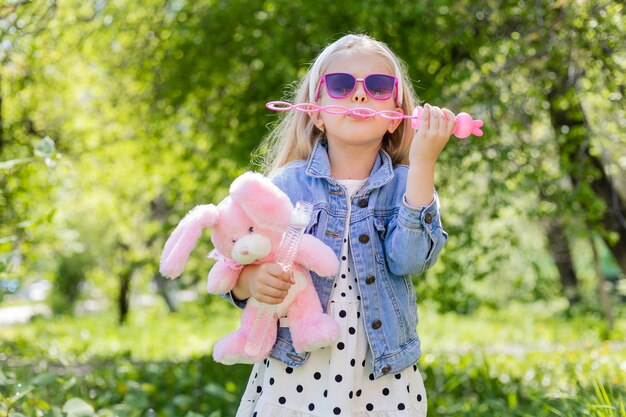 A happy little girl in summer with sunglasses inflates soap bubbles holds a toy in her hands