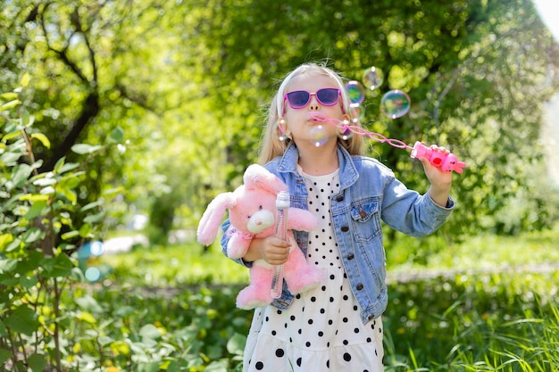 A happy little girl in summer with sunglasses inflates soap bubbles holds a toy in her hands