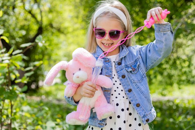 A happy little girl in summer with sunglasses inflates soap bubbles holds a toy in her hands