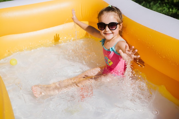膨脹可能なプールで水をはねかけるサングラスと水着の幸せな少女