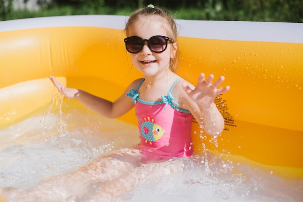 Happy little girl in sunglasses and swimsuit splashing water in the inflatable pool