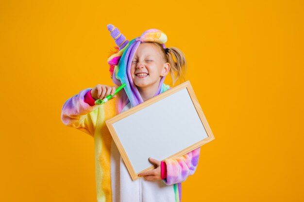 A happy little girl in a unicorn kigurumi holds an empty board for text on a yellow wall