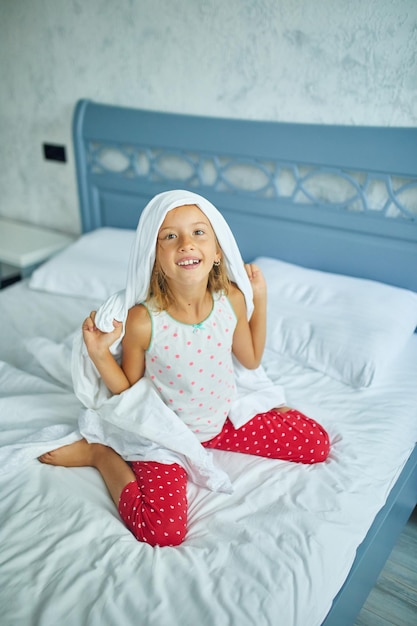 幸せな少女は、自宅の大きくて居心地の良いベッドの白いリネンで眠りから頭の上の楽しい毛布を目覚めさせます