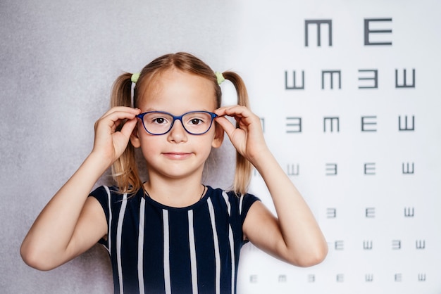 Happy little girl wearing glasses taking eyesight test before school with blurry eye chart at the background, child's vision examination