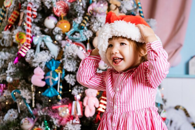 분홍색 드레스를 입고 크리스마스를 기다리는 동안 옆을 바라보는 산타 모자를 쓴 행복한 어린 소녀...