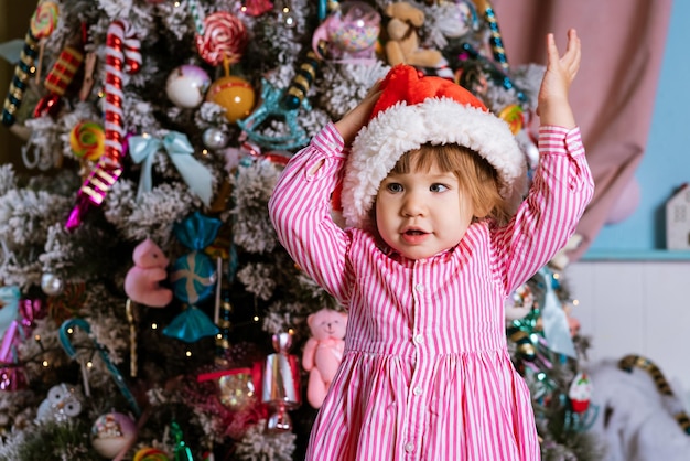 ピンクのドレスを着て、クリスマスを待っている間、横を向いているサンタの帽子をかぶっている幸せな少女...