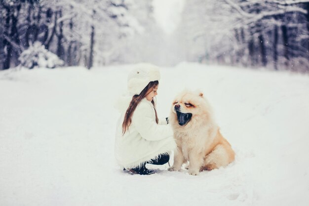 Happy little girl in white in hat sitting with dog in the winter park